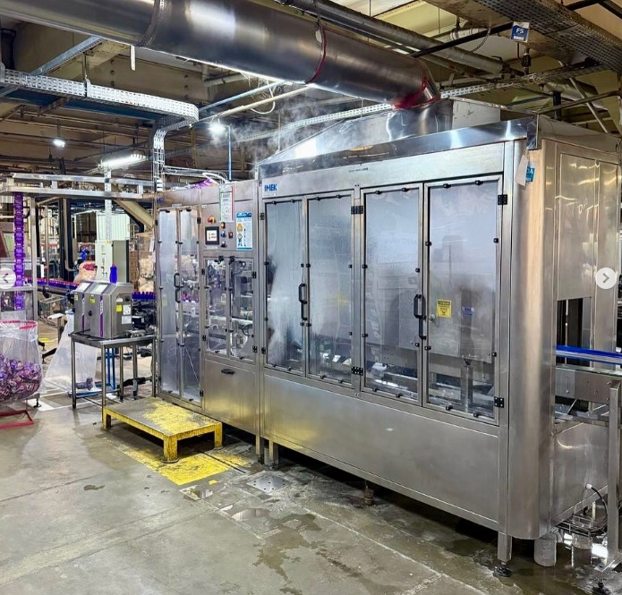 Rotuladora automática de sleeve IMEK aplicando rótulos termoencolhíveis em alta velocidade em linha de produção industrial moderna.