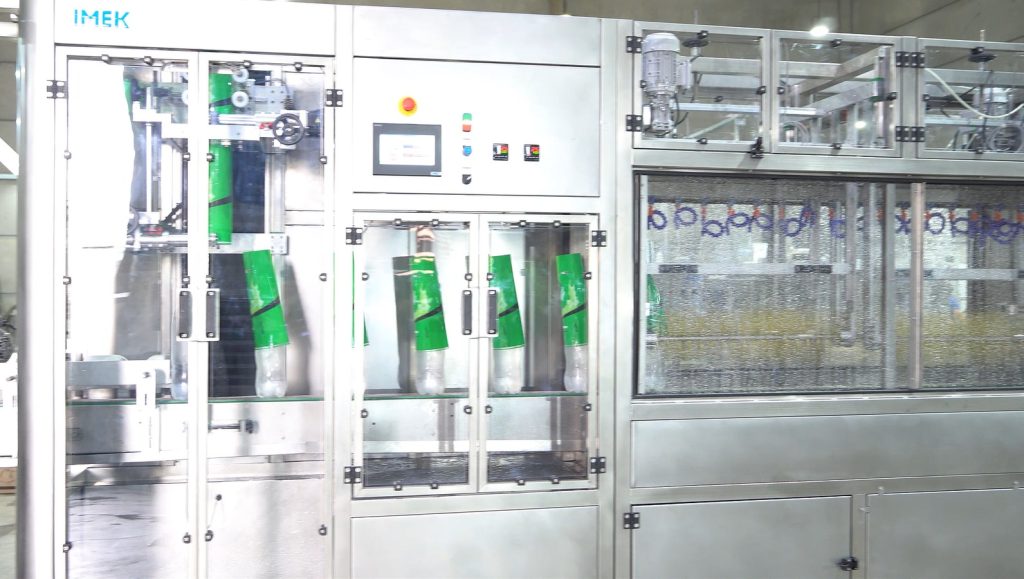 Rotuladora automática de sleeve IMEK aplicando rótulos termoencolhíveis em alta velocidade em linha de produção industrial moderna.